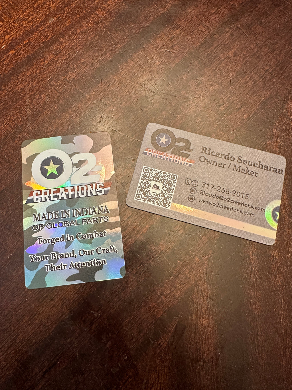 Two O2 Creations business cards on a dark brown wooden tabletop. One card features a camouflage pattern with a green star logo and text reading 'Made in Indiana of Global Parts' and 'Forged in Combat Your Brand, Our Craft, Their Attention.