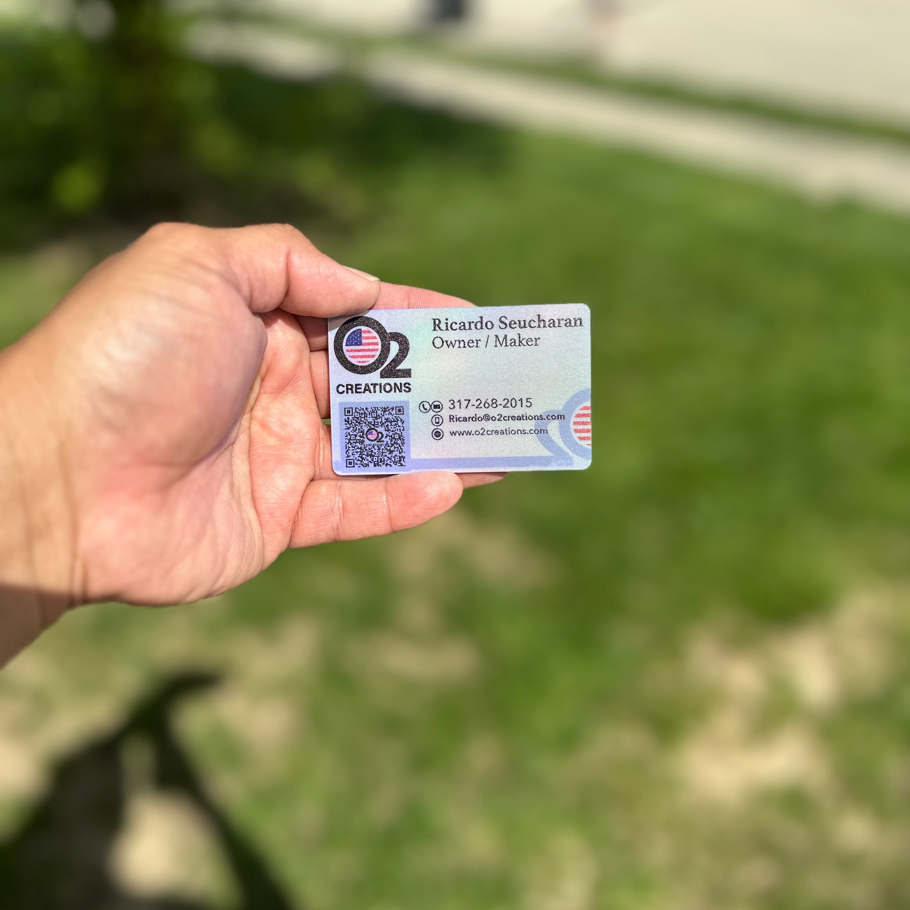 A hand holding a holographic business card with a QR code and contact information, outdoors.