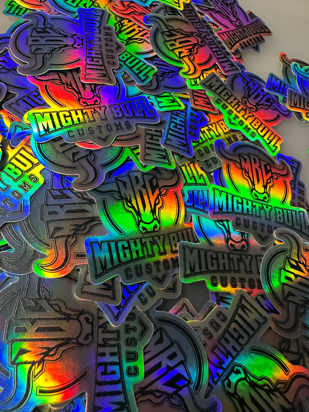 A pile of holographic die-cut stickers featuring a muscular bull head logo and the text 'Mighty Bull Custom' in green, reflecting rainbow colors.